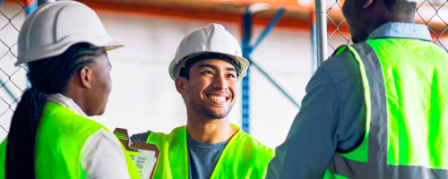 Three workers in safety vests and hard hats smiling in a warehouse