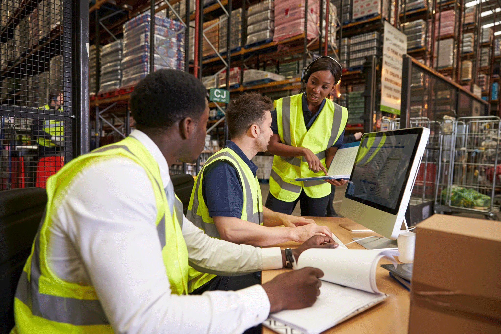 Three people in safety vests at a computer in a warehouse